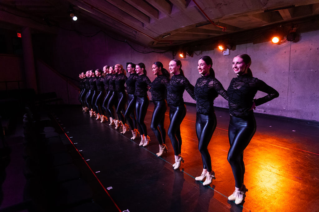 Dancers in a diagonal precision line under warm stage lighting