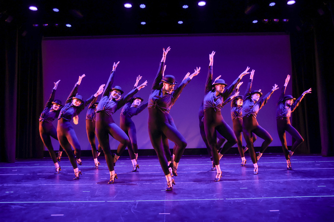 PDC dancers in a jazz formation with bowler hats, lit in purple stage lighting during a Performance Collective show